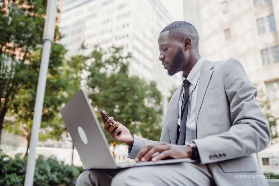 Businessman in a suit using a laptop and smartphone in an urban setting, blending technology with business.